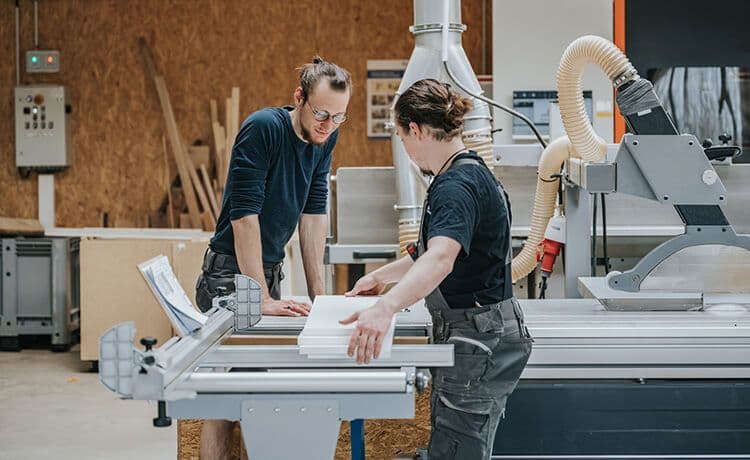 Zwei Zimmerer stehen in der Werkstatt und sägen ein Bauteil zurecht.