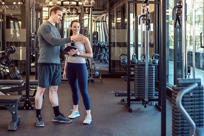 Fitnesstrainer zeigt einer jungen Frau ein Gerät im Fitnessstudio.