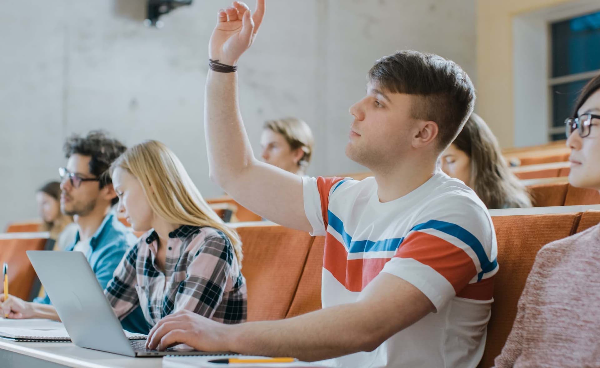 Student sitzt im Hörsaal mit anderen Studierenden und Laptop und zeigt auf