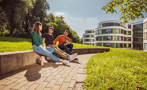 Ansicht eines Gebäudes und drei Personen sitzen davor auf einer Wiese.