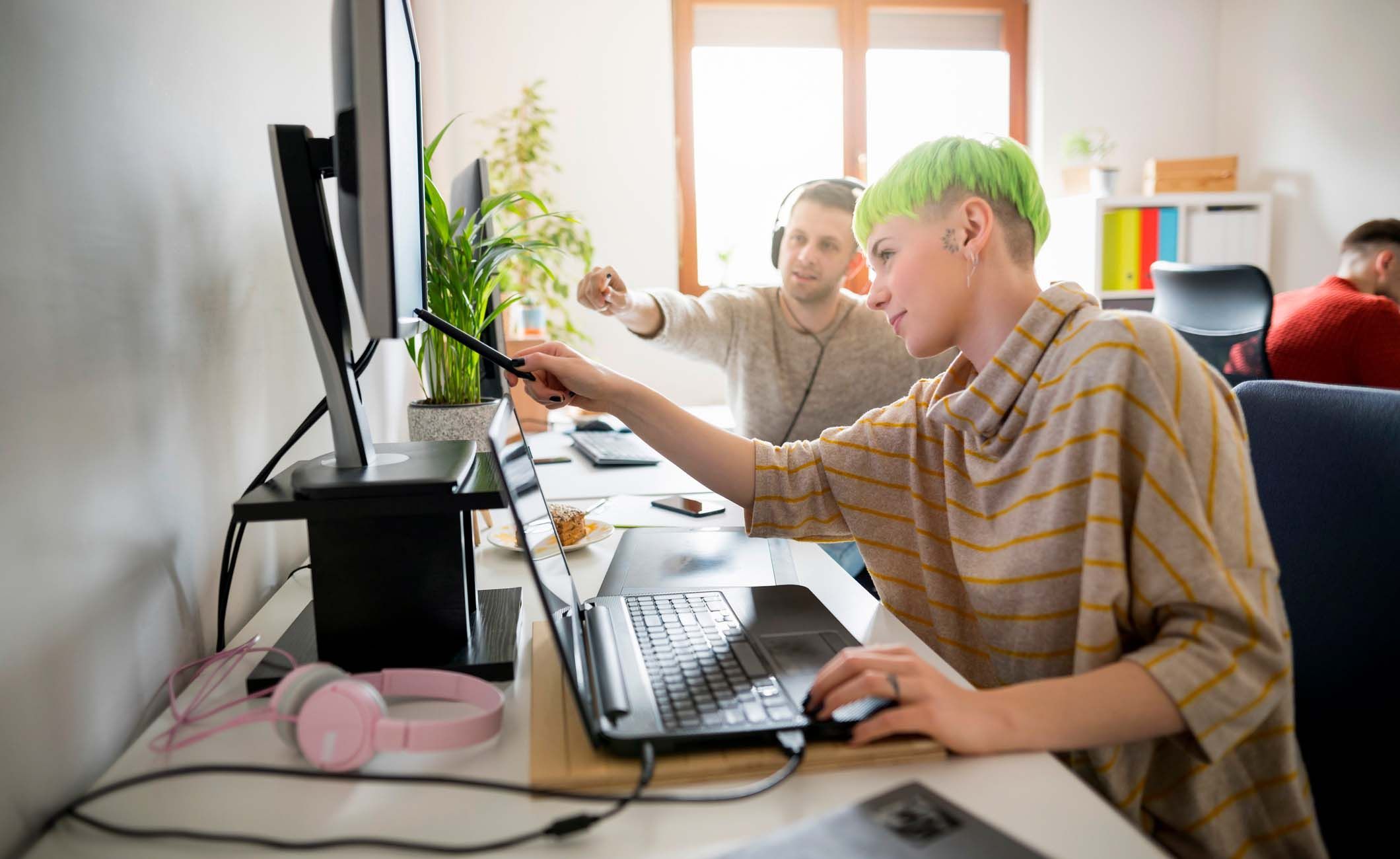 Eine Frau mit kurzen, grünen Haaren und ein Mann mit Kopfhörern zeigen auf einen Bildschirm.