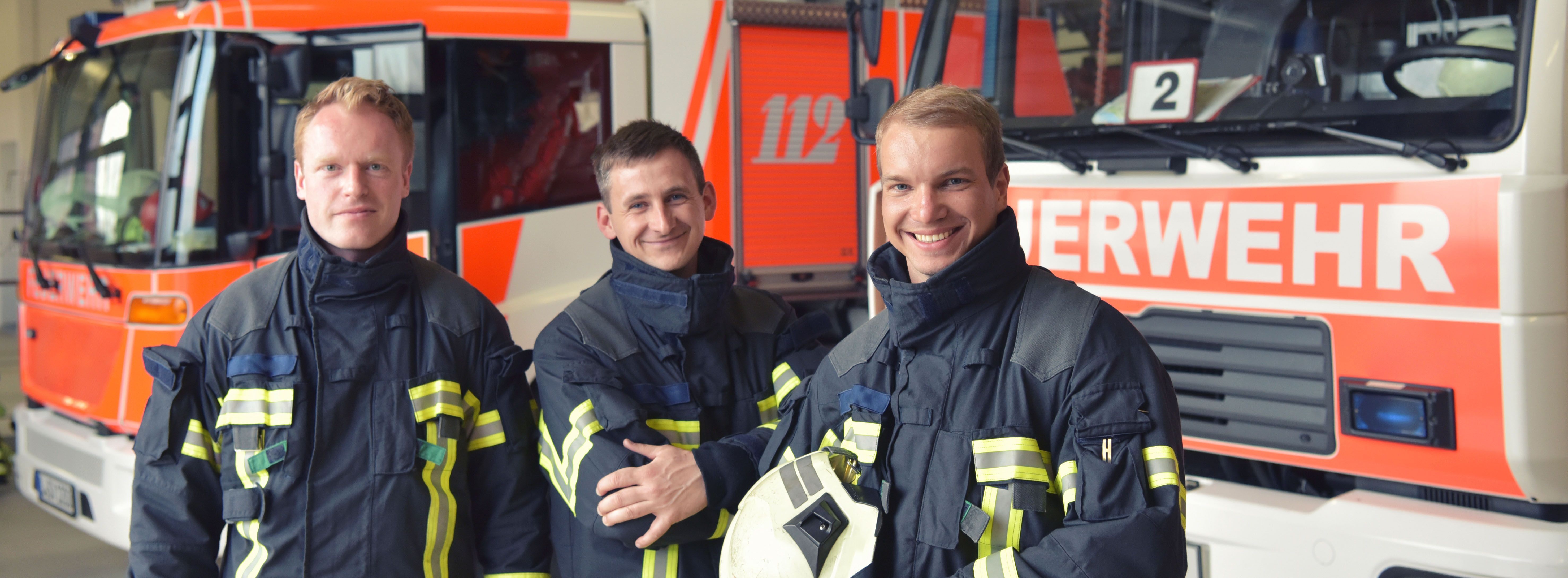 Drei Feuerwehrmänner in Ausrüstung stehen vor Feuerwehr-Autos und lächeln in die Kamera.