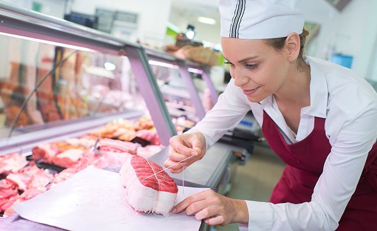 Junge Frau mit Schürze verpackt an der Fleischtheke ein Stück Fleisch.