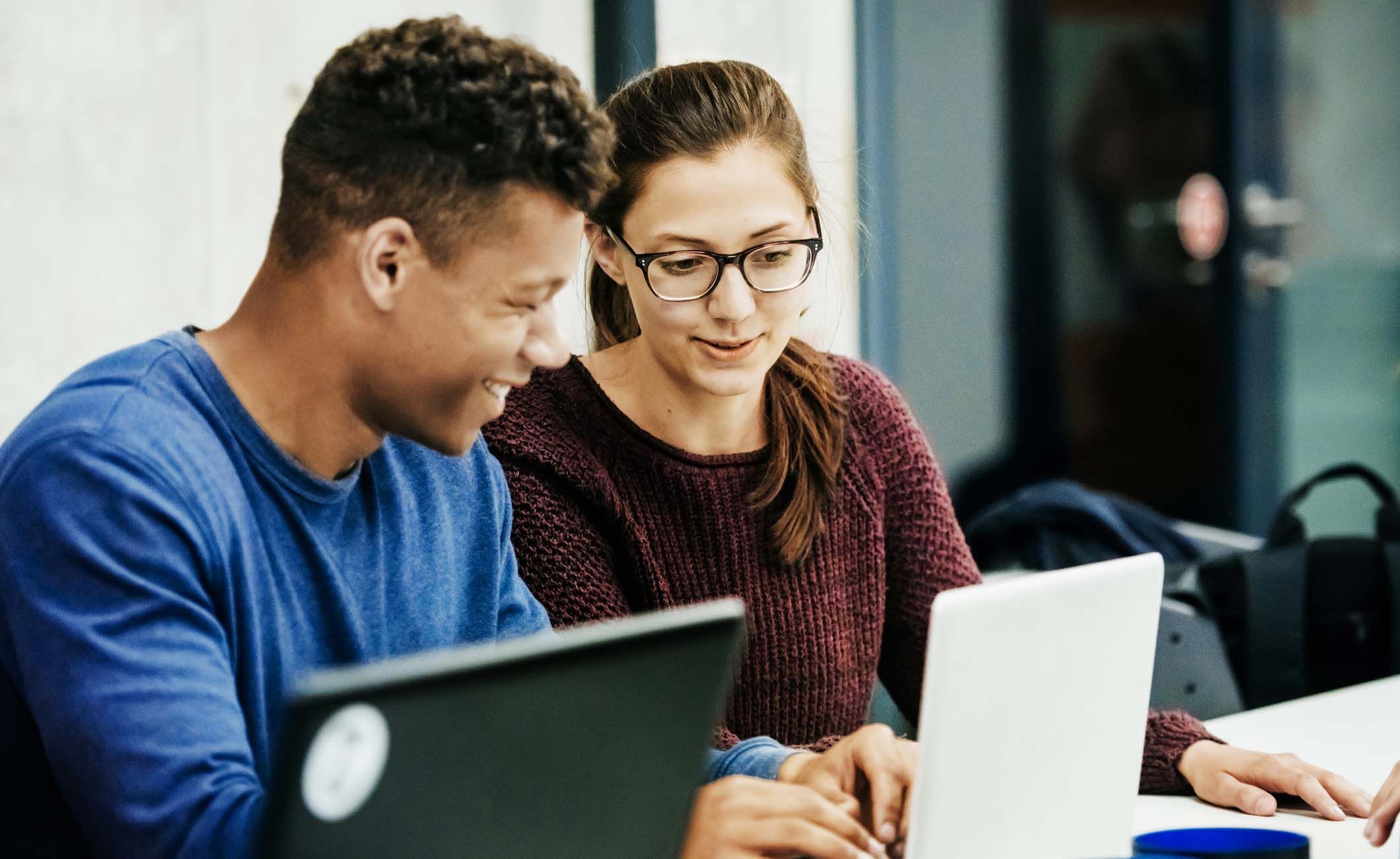 Junge Frau mit Brille und ein Mann sitzen vor Laptops und diskutieren