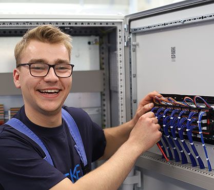 Azubi vor einem elektrischen Schrank lacht in die Kamera.