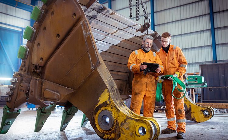 Ein Ausbilder zeigt einem Azubi etwas in einer Halle neben einem Baugerät. Ein Ausbilder zeigt einem Azubi etwas in einer Halle neben einem Baugerät.