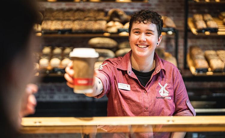 Frau in der Bäckerei reicht einem Kunden einen Becker Kaffee.