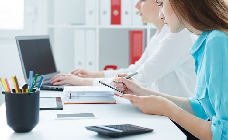 Zwei junge Frauen sitzen an einem Schreibtisch mit Laptop, Stiften, Taschenrechner und Tablet.