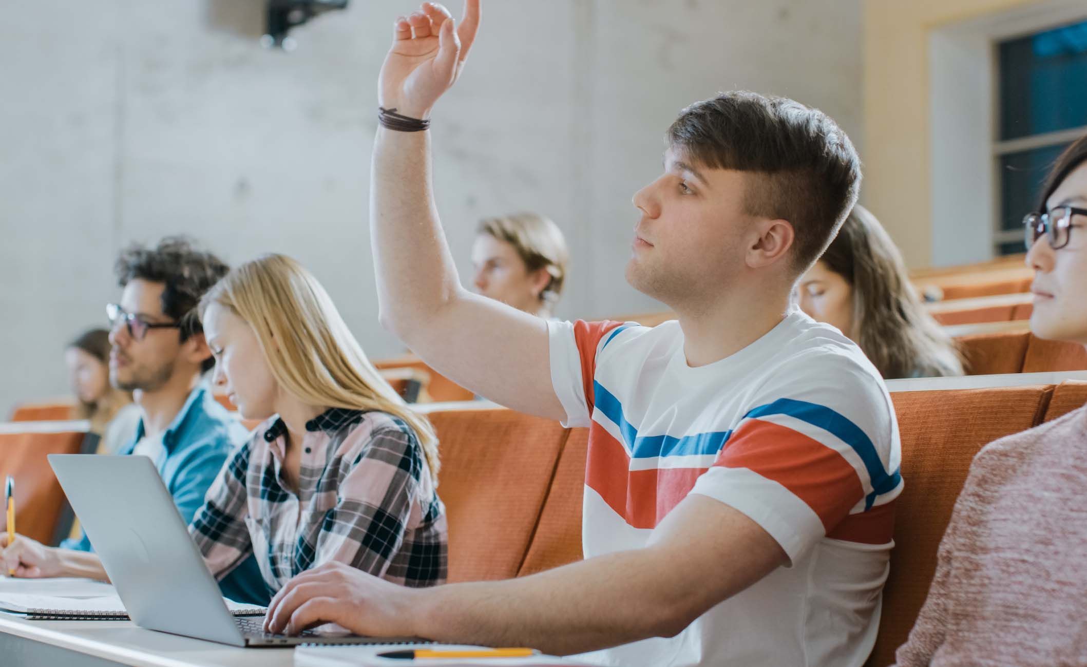 Student sitzt im Hörsaal mit anderen Studierenden und Laptop und zeigt auf
