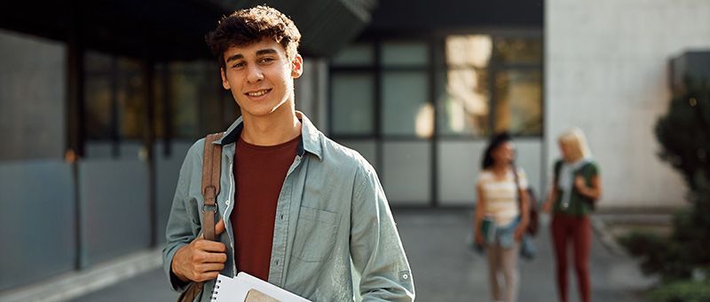 Junger Schüler steht lächelnd vor einer Fachoberschule. Junger Schüler steht lächelnd vor einer Fachoberschule.