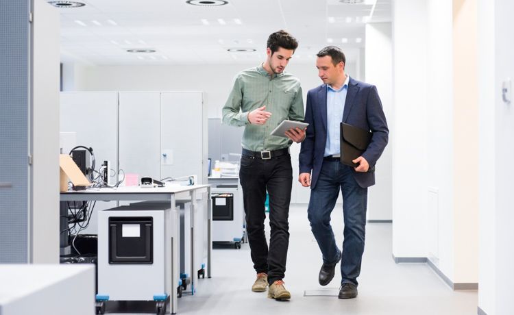 Zwei männliche Finanzbeamte unterhalten sich, während sie einen Büroflur entlang gehen.