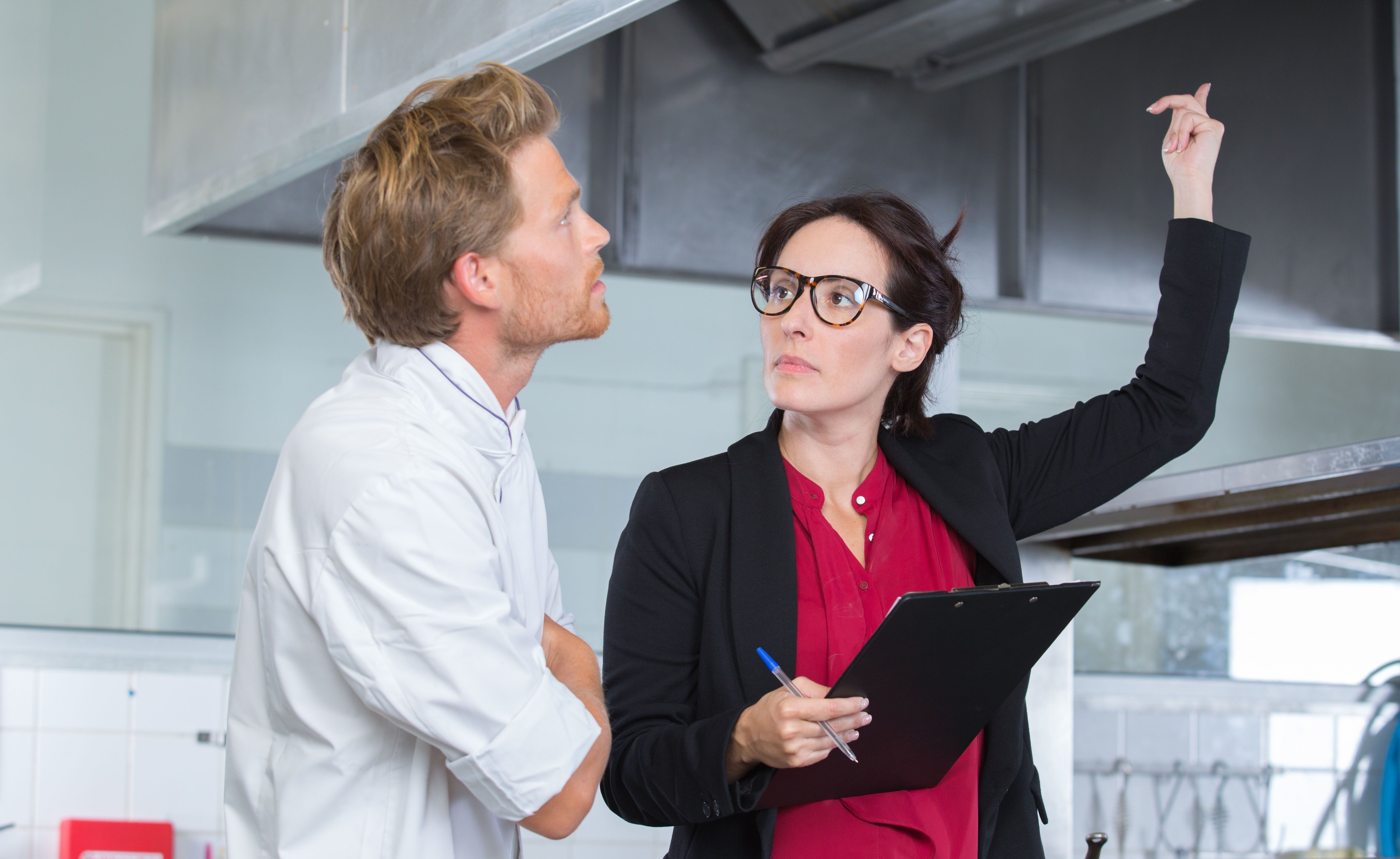 Inspektorin prüft Restaurantküche mit jungem Koch.