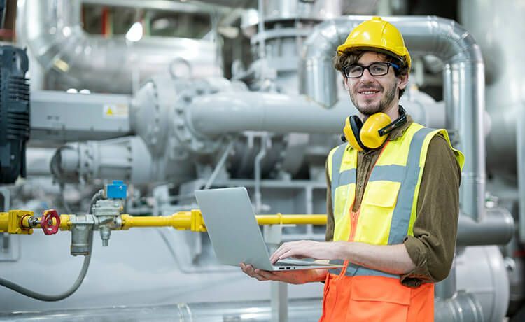 Mechatroniker für Kältetechnik steht in einer Industriehalle mit Rohren und hält einen Laptop in der Hand.