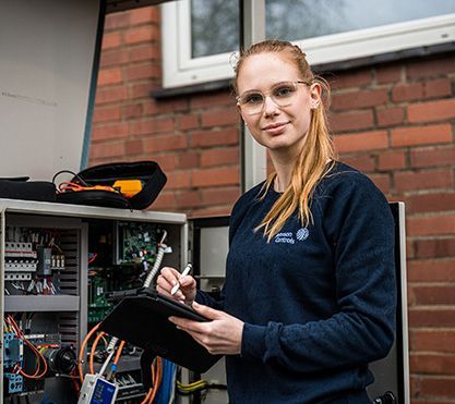 Junge Frau steht vor einem Kasten mit Kabel und schreibt etwas auf.