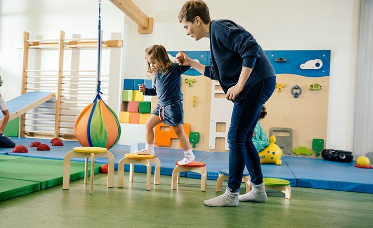Frau hilft einem Kind beim Balancieren auf Stühlen.