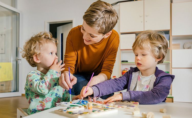 Frau hilft zwei Kindern beim Malen mit Wasserfarben.