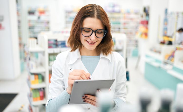 Junge PTA mit Tablet hin der Hand arbeitet in einer Apotheke.