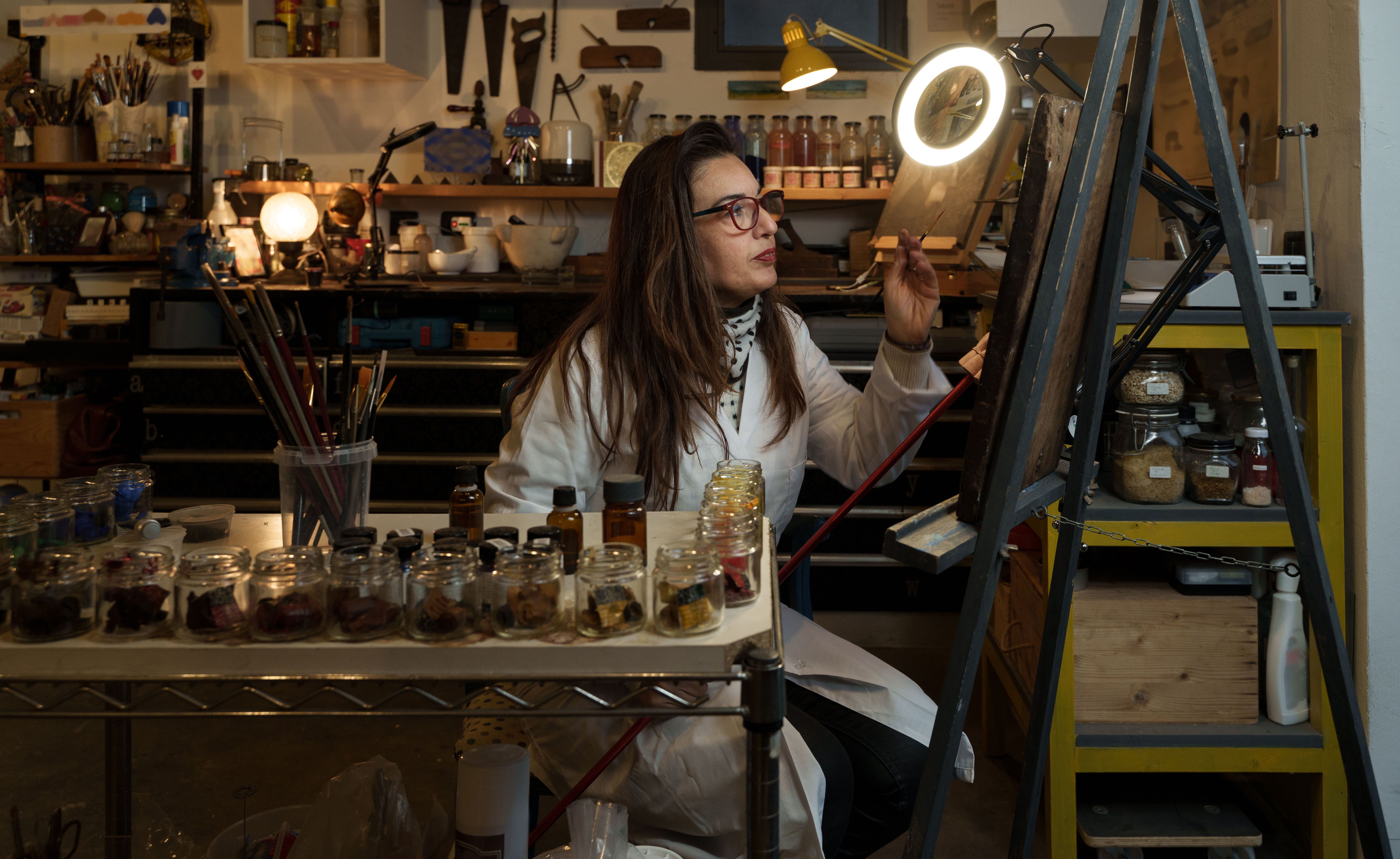 Eine Restauratorin restauriert ein antikes Gemälde in ihrem Atelier und nimmt mit einem Pinsel Ausbesserungen vor. Im Hintergrund befindet sich ein Regal, das mit Farben und Werkzeugen gefüllt ist. Eine Restauratorin restauriert ein antikes Gemälde in ihrem Atelier und nimmt mit einem Pinsel Ausbesserungen vor. Im Hintergrund befindet sich ein Regal, das mit Farben und Werkzeugen gefüllt ist.