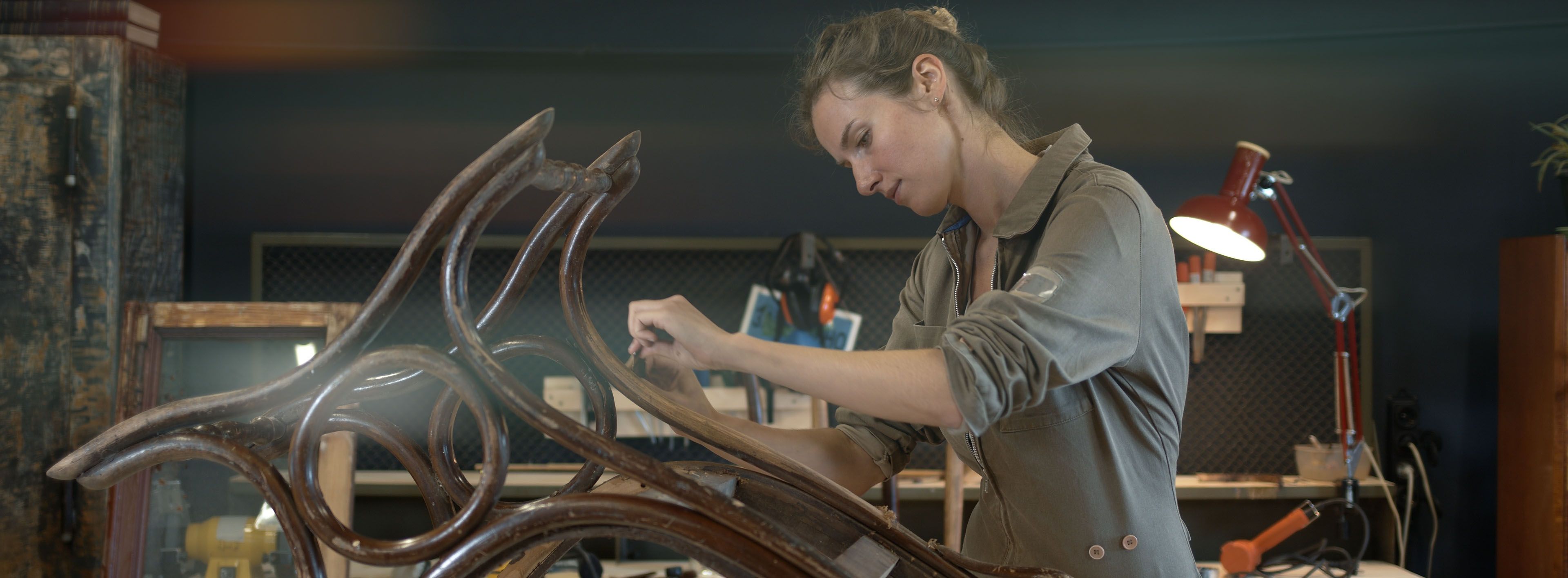 Restaurierung eines Holzschaukelstuhls in einer Werkstatt. Eine Holzrestauratorin entfernt mit einem Spachtel Farbe von den Beinen des Schaukelstuhls.