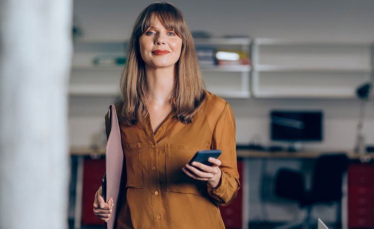 Junge Social Medie Managerin hält ein Smartphone und Unterlagen in den Händen und lächelt in die Kamera.