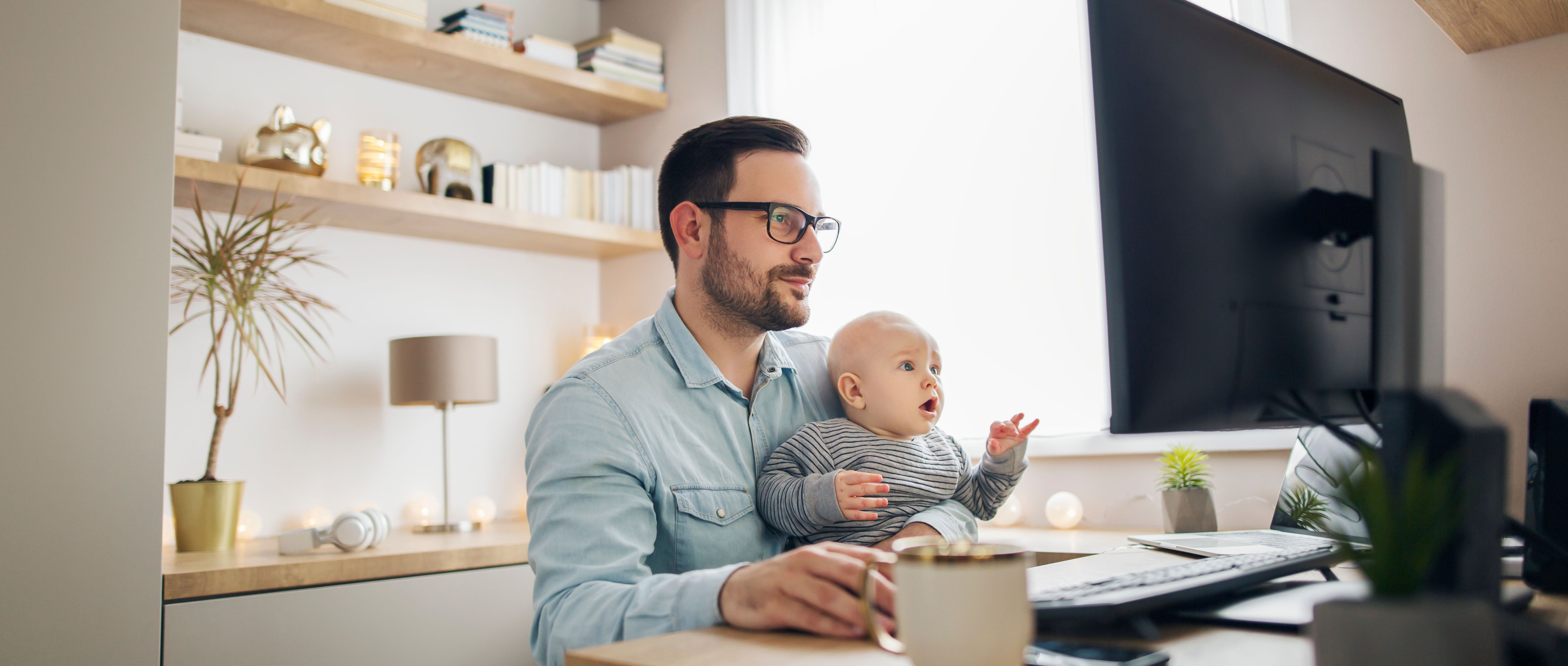 Vater sitzt mit Baby im Home Office am PC.