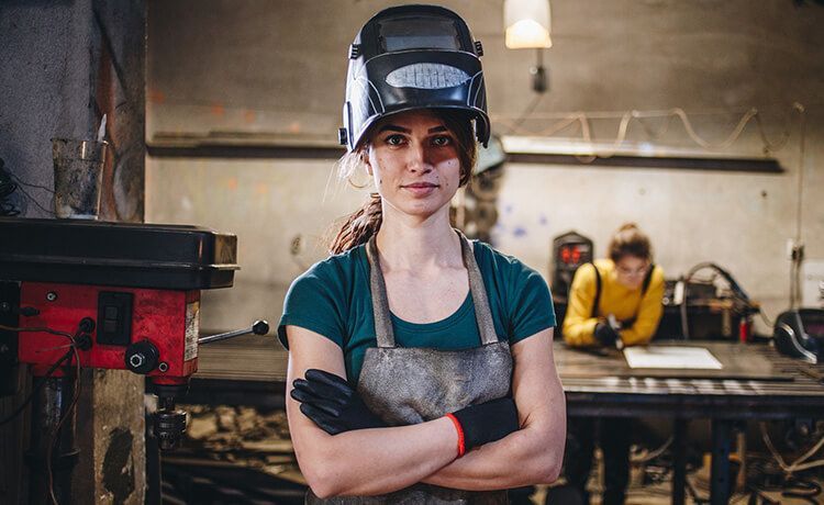 Junge Schweißerin steht mit hochgezogener Schweißer-Maske und verschränkten Armen in der Werkstatt und schaut in die Kamera.