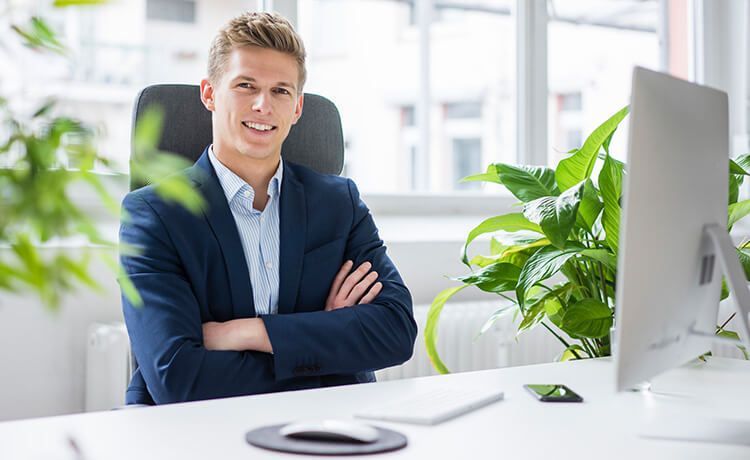 Junger Wirtschaftsprüfer im Anzug sitzt am Schreibtisch und lächelt. Junger Wirtschaftsprüfer im Anzug sitzt am Schreibtisch und lächelt.