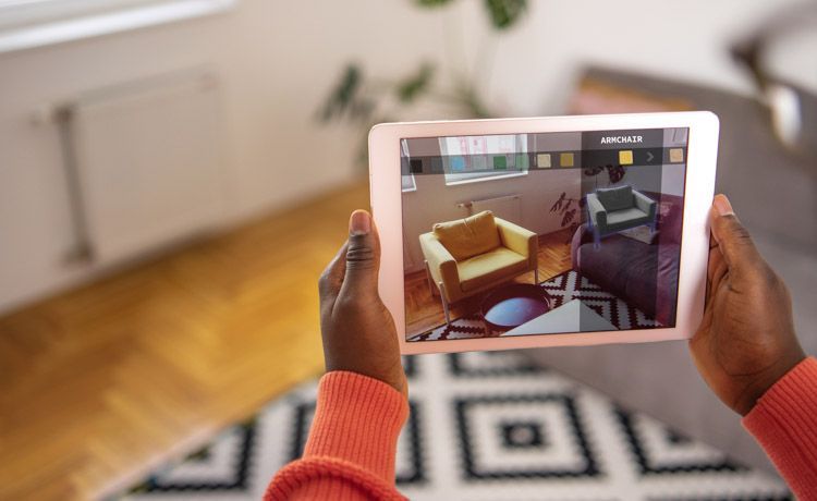 Eine Frau hält ein Tablet in den Händen auf denen Möbel zu sehen sind, die mithilfe von Augmented Reality so aussehen, als würden sie in ihrem Wohnzimmer stehen.