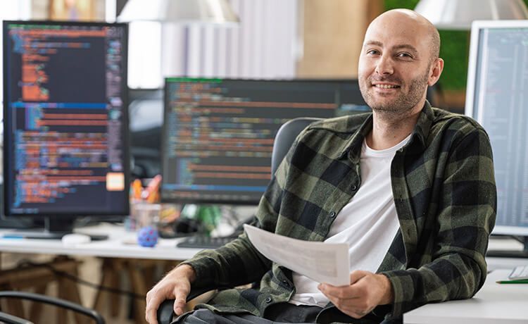 Programmierer sitzt vor einem Computer mit Code und lächelt in die Kamera.