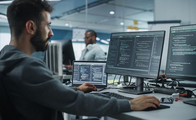 Junger Informatik-Student sitzt vor mehreren Bildschirmen mit Quellcode.