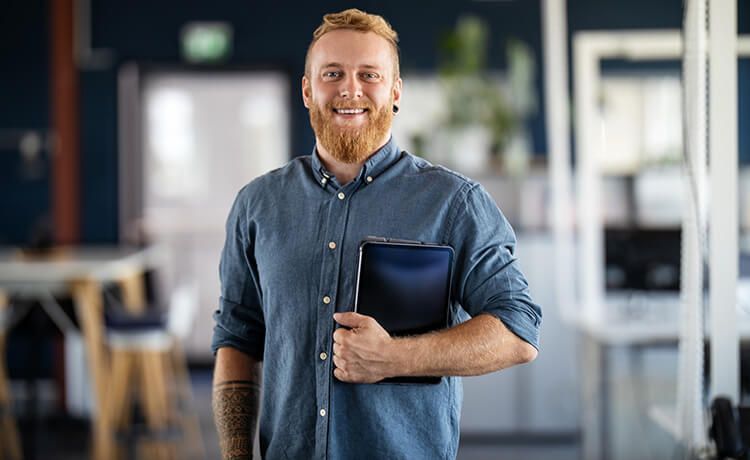 Informatik-Student mit Bart hält Tablet in der Hand und lächelt.