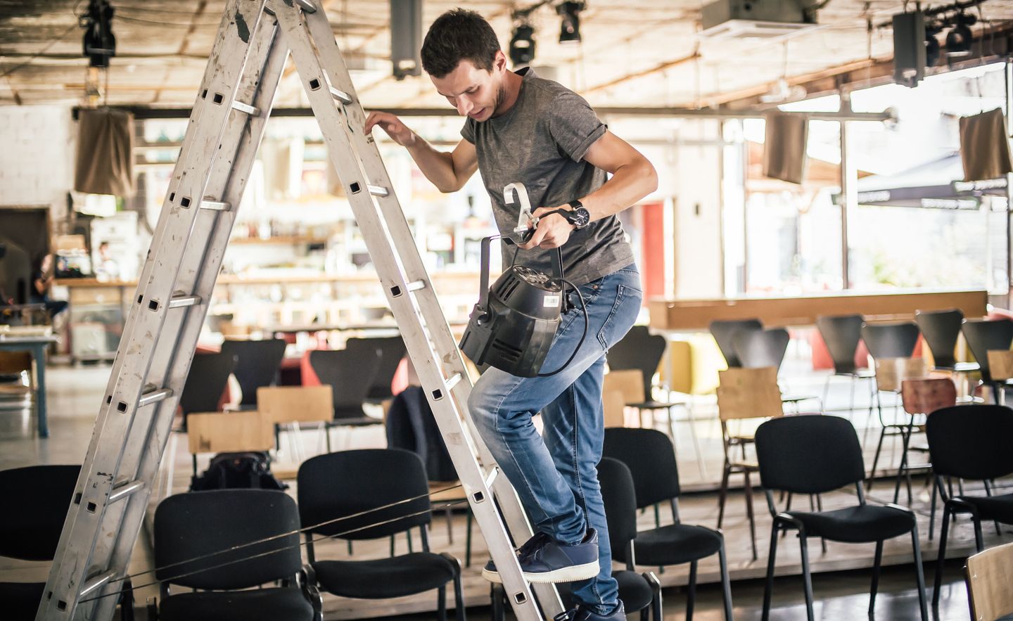 Veranstaltungstechniker hängt Lampen für eine Theaterbühne auf einer Leiter auf.