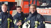 Drei Feuerwehrmänner in Ausrüstung stehen vor Feuerwehr-Autos und lächeln in die Kamera.