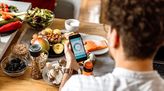 Junger Ernährungsberater sitzt an einem Tisch voller gesunder Lebensmittel und checkt eine Kalorientracker-App am Smartphone.