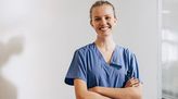 Krankenschwester steht vor einer Wand im Krankenhaus und blickt lächelnd in die Kamera.