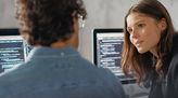 Zwei junge Programmierer sitzen vor Computer mit Code und unterhalten sich über das Programmieren.