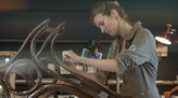 Restaurierung eines Holzschaukelstuhls in einer Werkstatt. Eine Holzrestauratorin entfernt mit einem Spachtel Farbe von den Beinen des Schaukelstuhls.